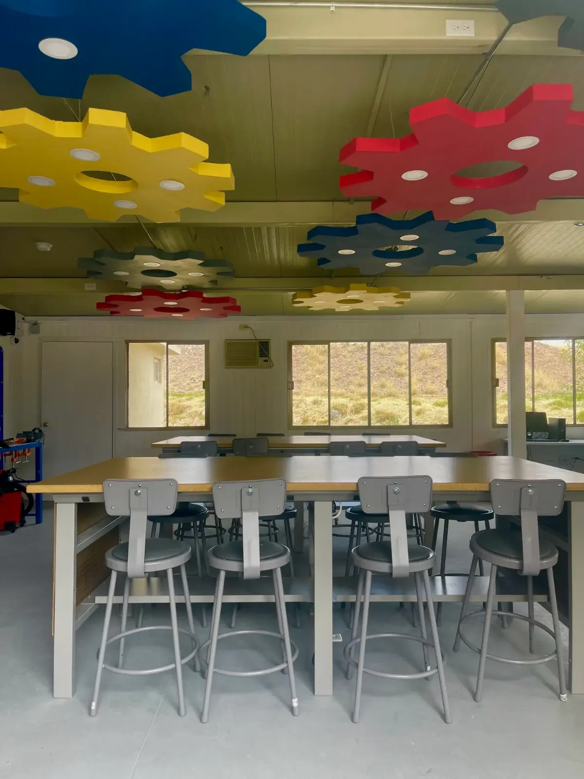 Aula moderna con techo de formas geométricas coloridas, escritorios altos y sillas para estudiantes, ventanales con vista al exterior
